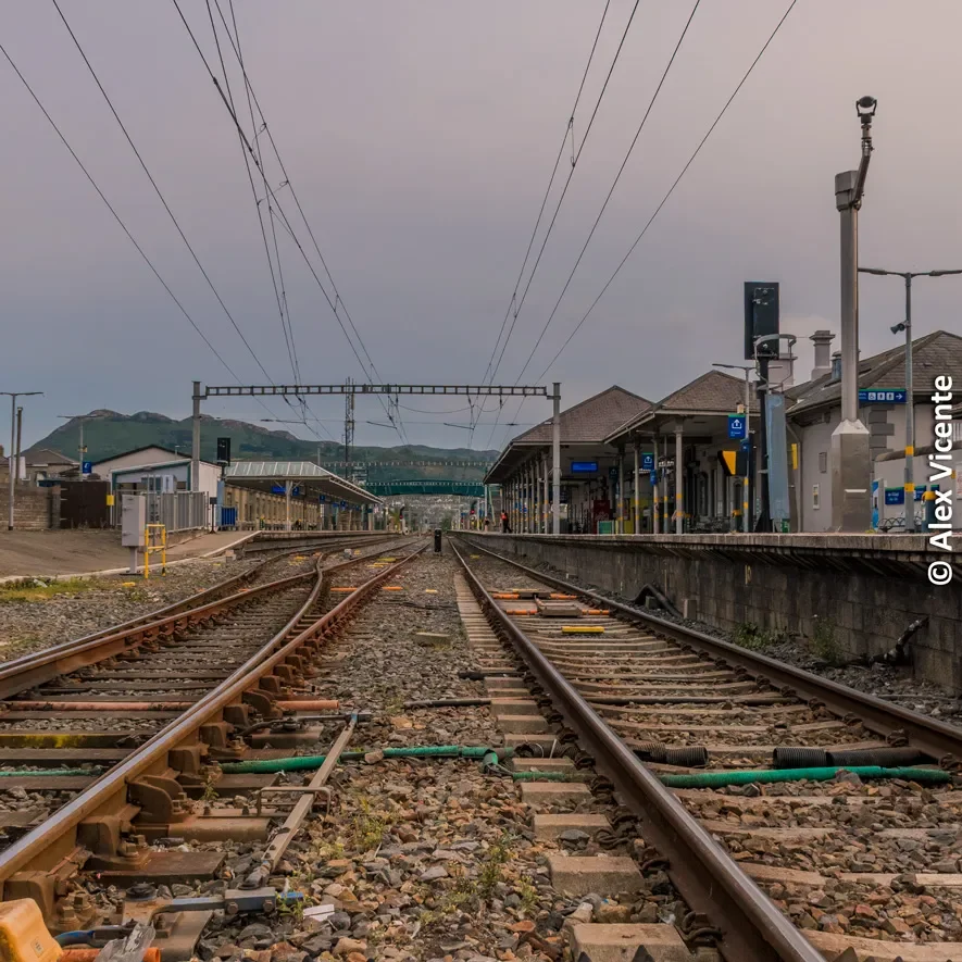 Railway tracks leading from a quiet station towards mountains.